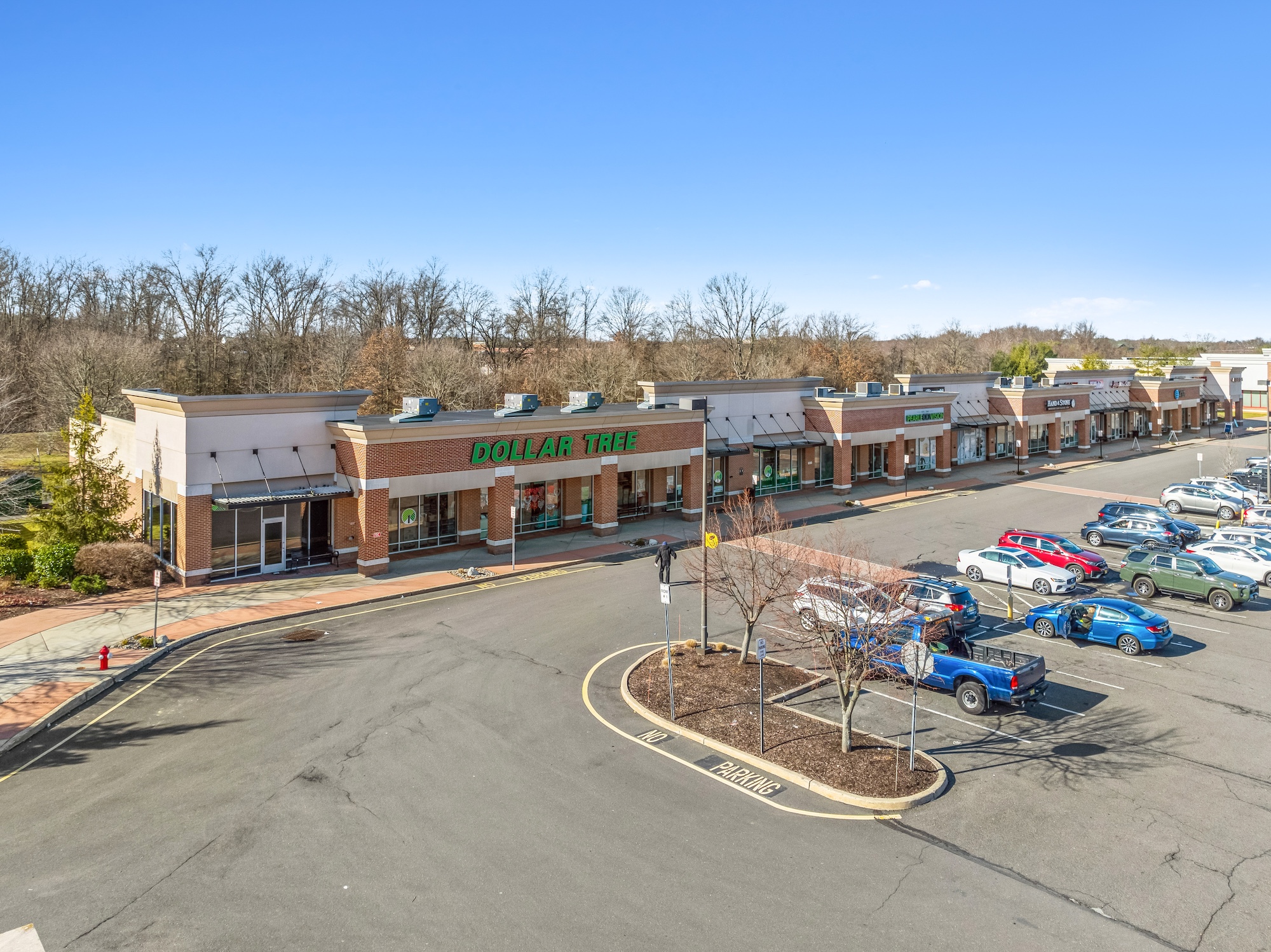 Aerial photograph of an inline retail storefront strip with tenant signage, parking, and landscaping