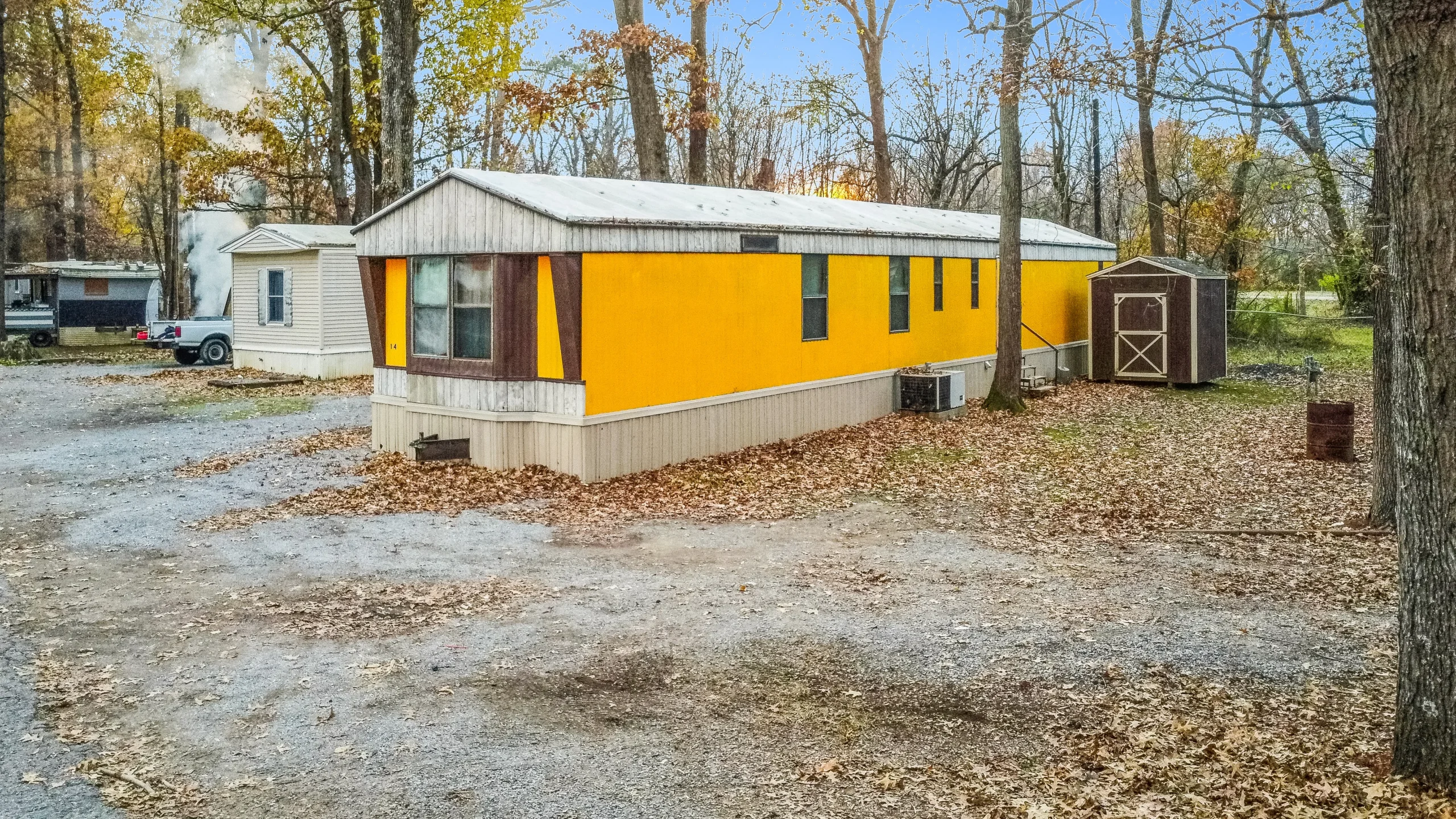 Ground-level photography of single-wide manufactured home with storage shed in rural wooded setting