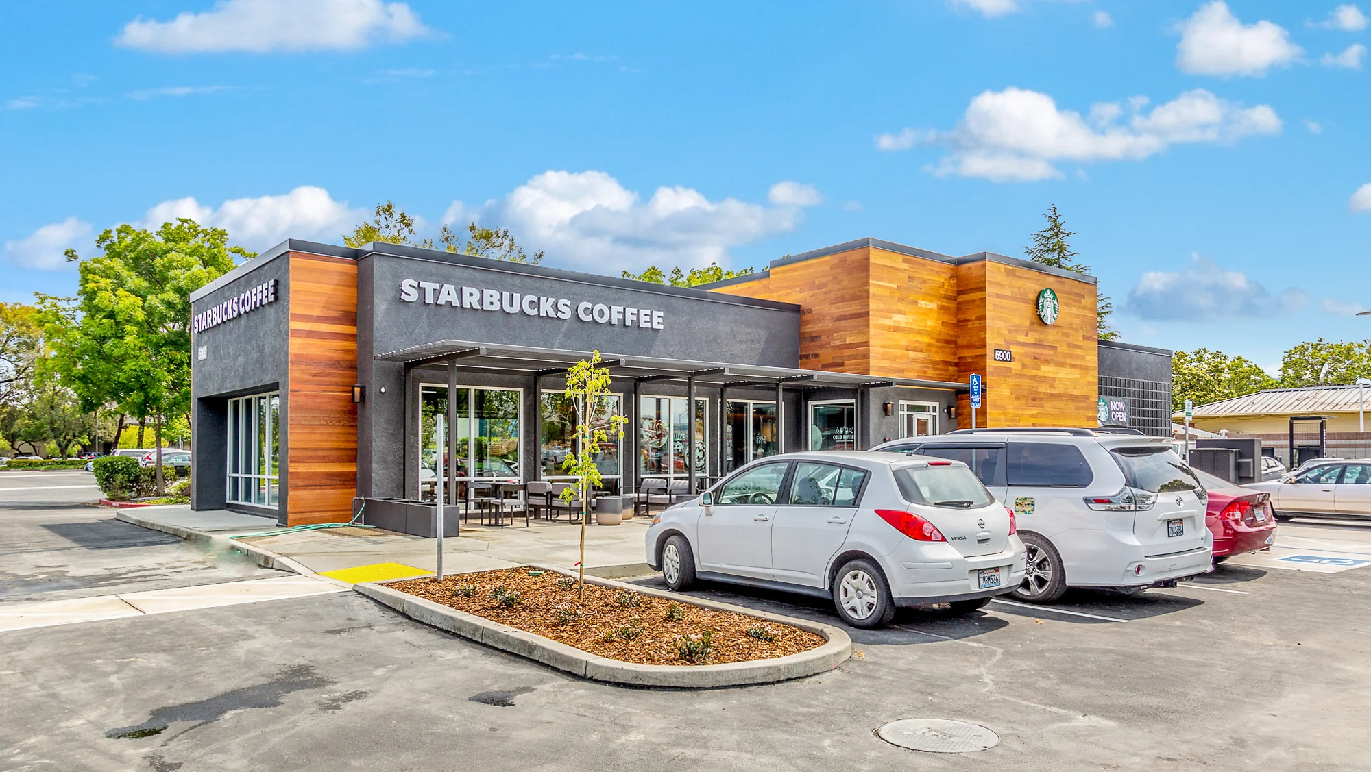 Ground-level photograph of a single-tenant Starbucks Coffee shop with modern wood and stucco facade, outdoor patio seating, and customer parking
