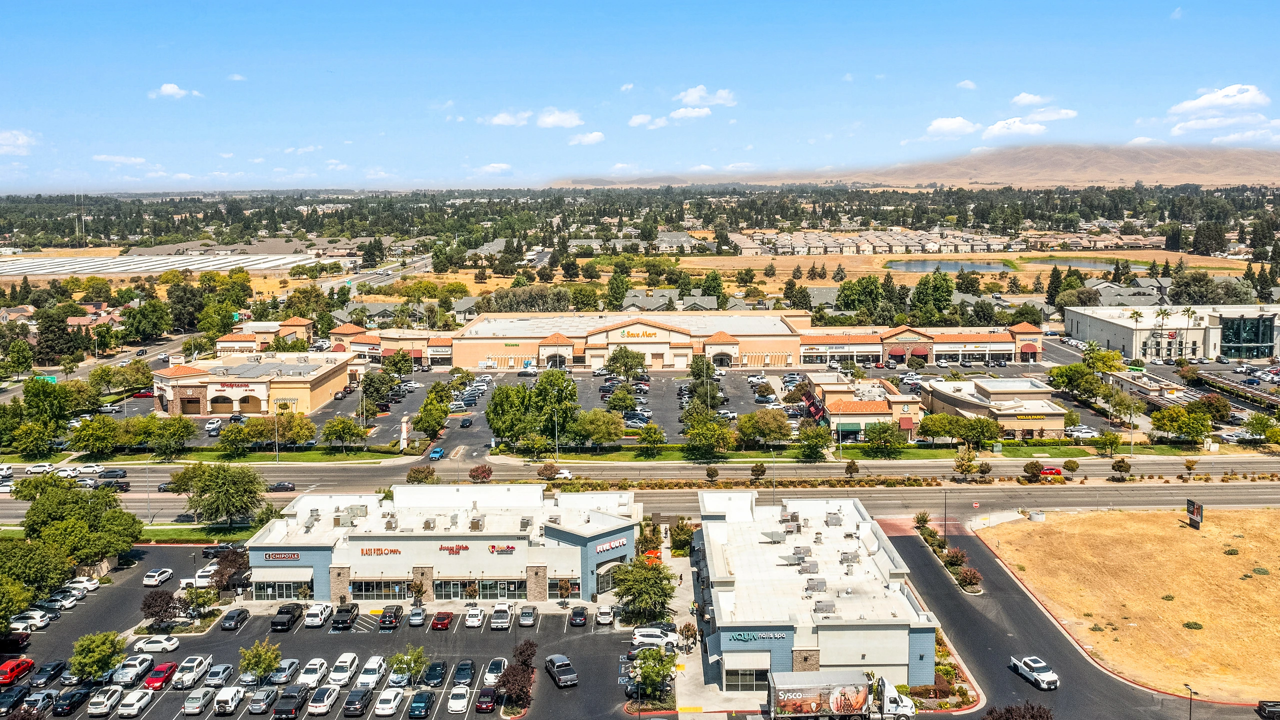 Aerial photograph of a power center with tile-roof large-format retail buildings, restaurant pads, and a second retail strip across the arterial road