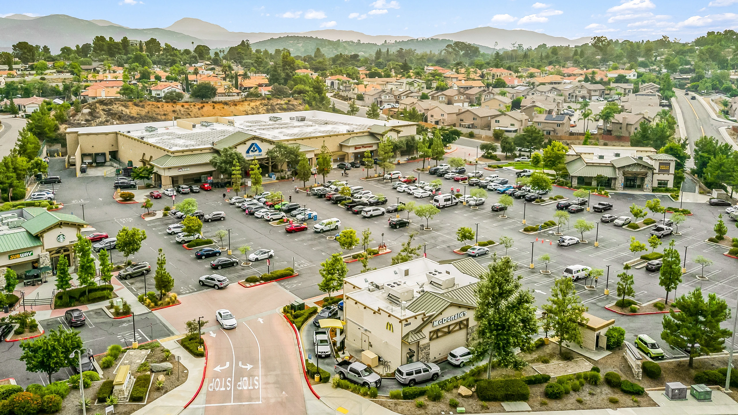 Aerial photograph of a grocery-anchored shopping center with pharmacy, bank and restaurant outparcel pads, deep landscaped parking field, and surrounding residential neighborhood
