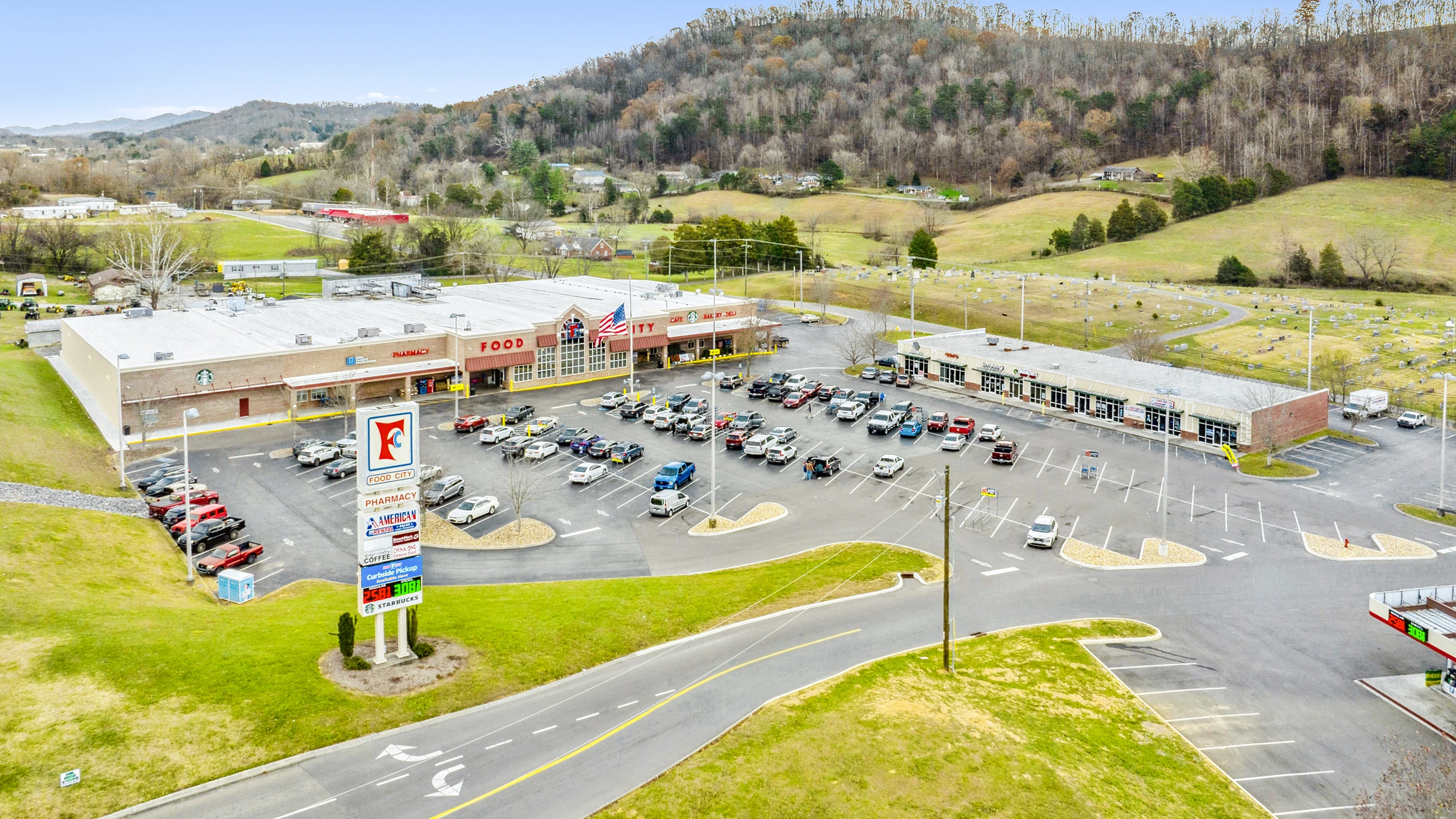 Aerial photograph of a grocery-anchored shopping center with attached pharmacy, cafe, inline retail strip, and pylon signage in a rural mountain trade area