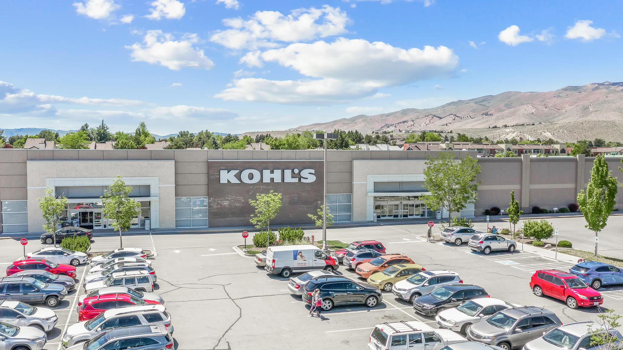 Aerial photograph of a single-tenant Kohl's department store with dual customer entrances, full parking field, and mountain backdrop