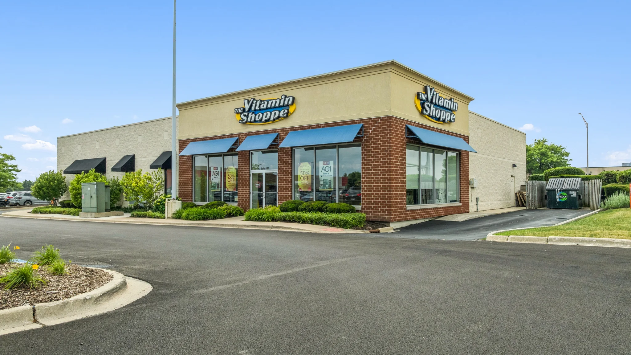 Ground-level photograph of a single-tenant Vitamin Shoppe retail storefront with corner entry, brick facade, awnings, and drive aisle